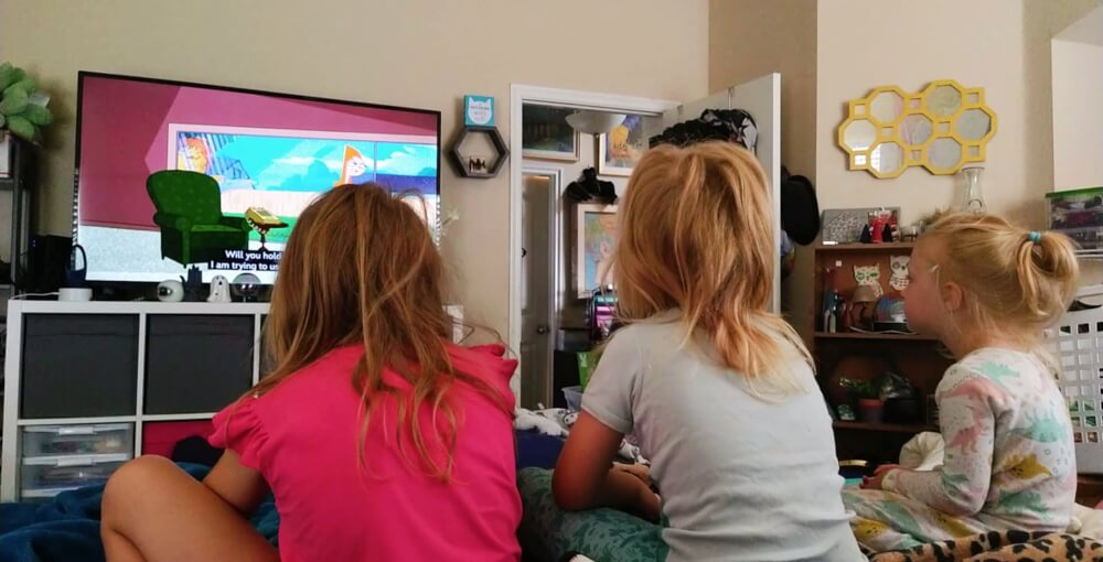 Three girls, from oldest to youngest, sitting on a bed watching Phineas & Ferb